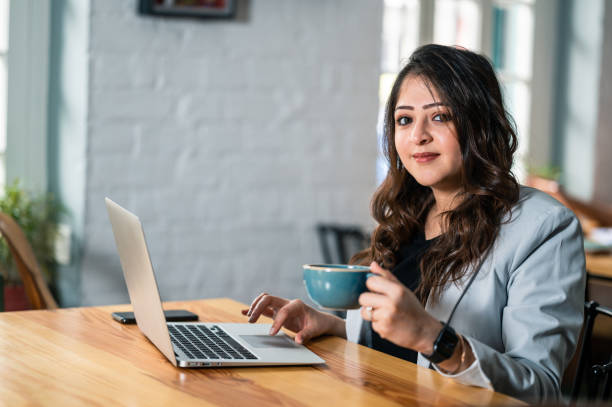 Indoor image of young Asian/Indian Business woman at coffee shop working using laptop and drinking coffee or tea in blue cup.